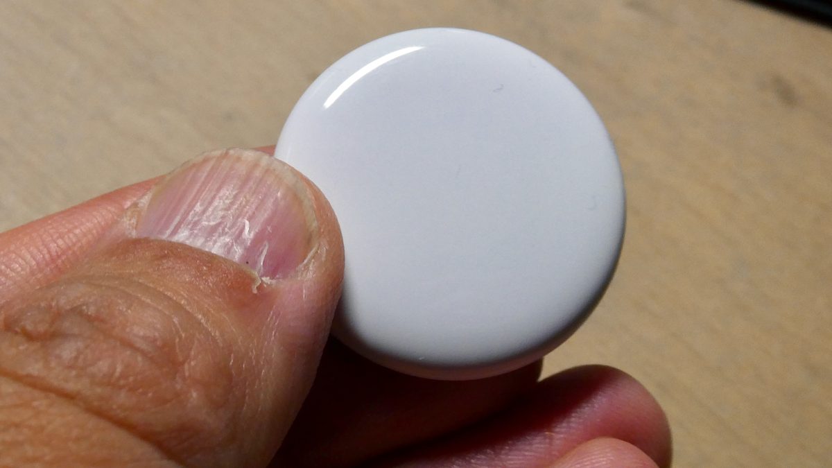 Mão segurando um pequeno rastreador Bluetooth redondo e branco sobre uma mesa de madeira