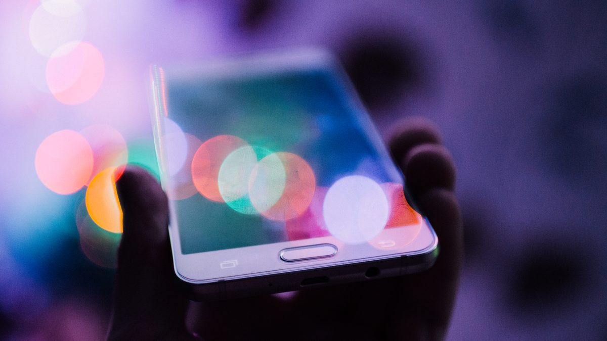 Mão segurando um celular com a tela acesa refletindo luzes coloridas desfocadas ao fundo