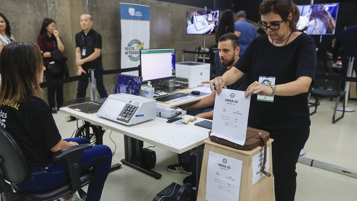 Mulher segura uma folha com a identificação Urna 33 ao lado da urna eletrônica, durante o teste de integridade de urnas em São Paulo, neste domingo (4)