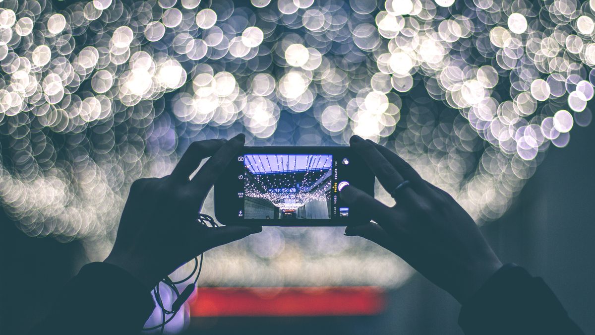 Duas mãos seguram um celular na horizontal filmando luzes desfocadas, com a câmera aberta na tela