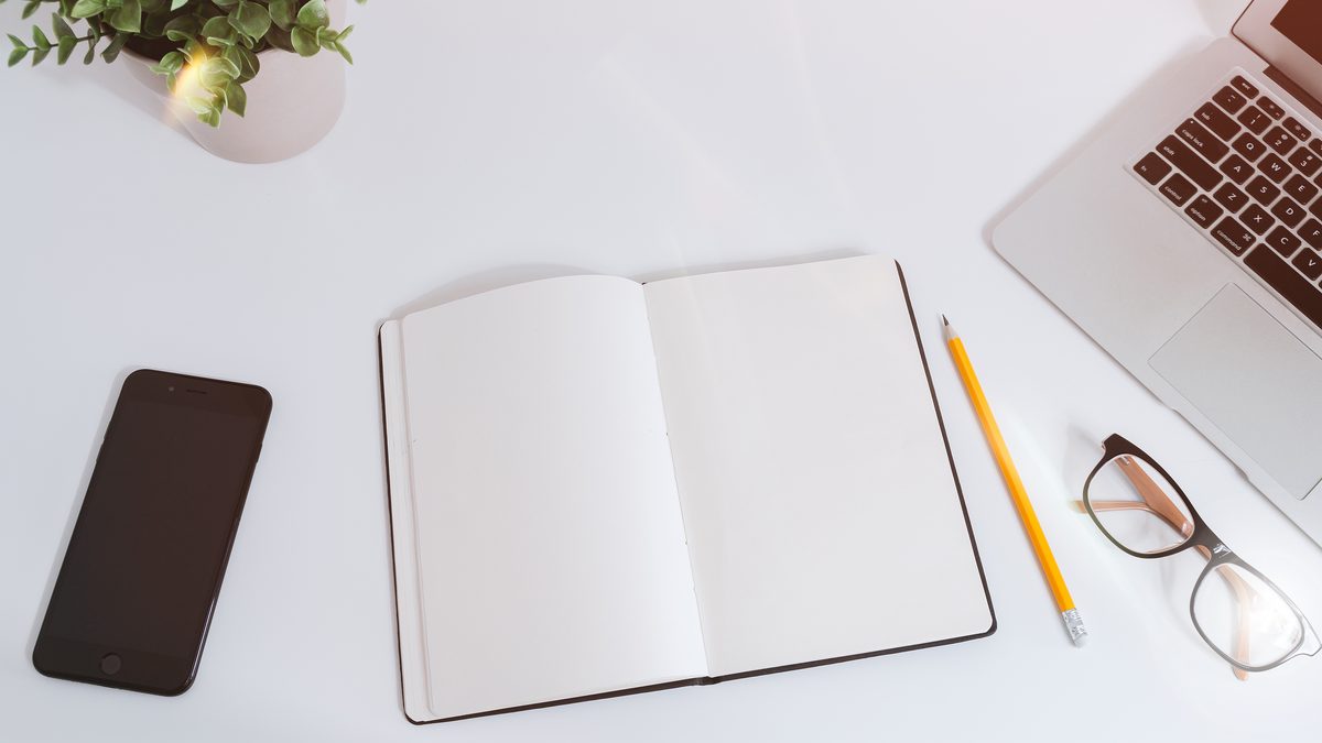 Mesa de trabalho branca com celular de tela apagada, caderno aberto, lápis, óculos e notebook