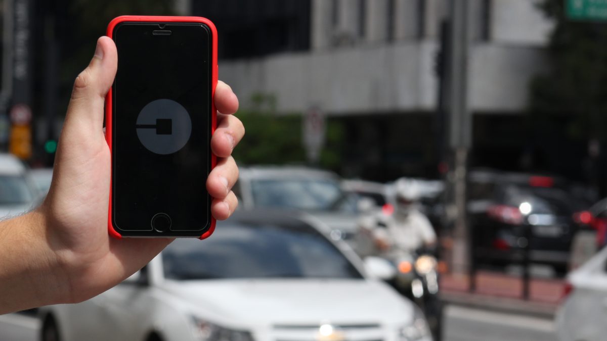 Mão segurando um celular com o aplicativo da Uber aberto, com carros passando ao fundo numa avenida de São Paulo