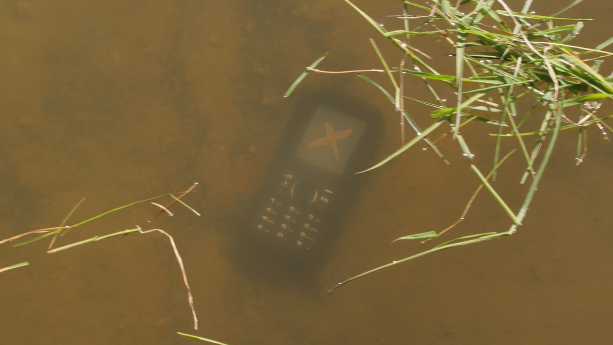 Celular submerso em água turva, com folhas de capim boiando por cima