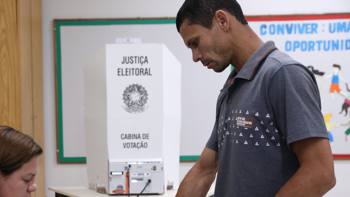 Eleitor toca o leitor de digital do terminal da mesa receptora, com a cabine de votação da Justiça Eleitoral ao fundo, no 2º turno de 2022