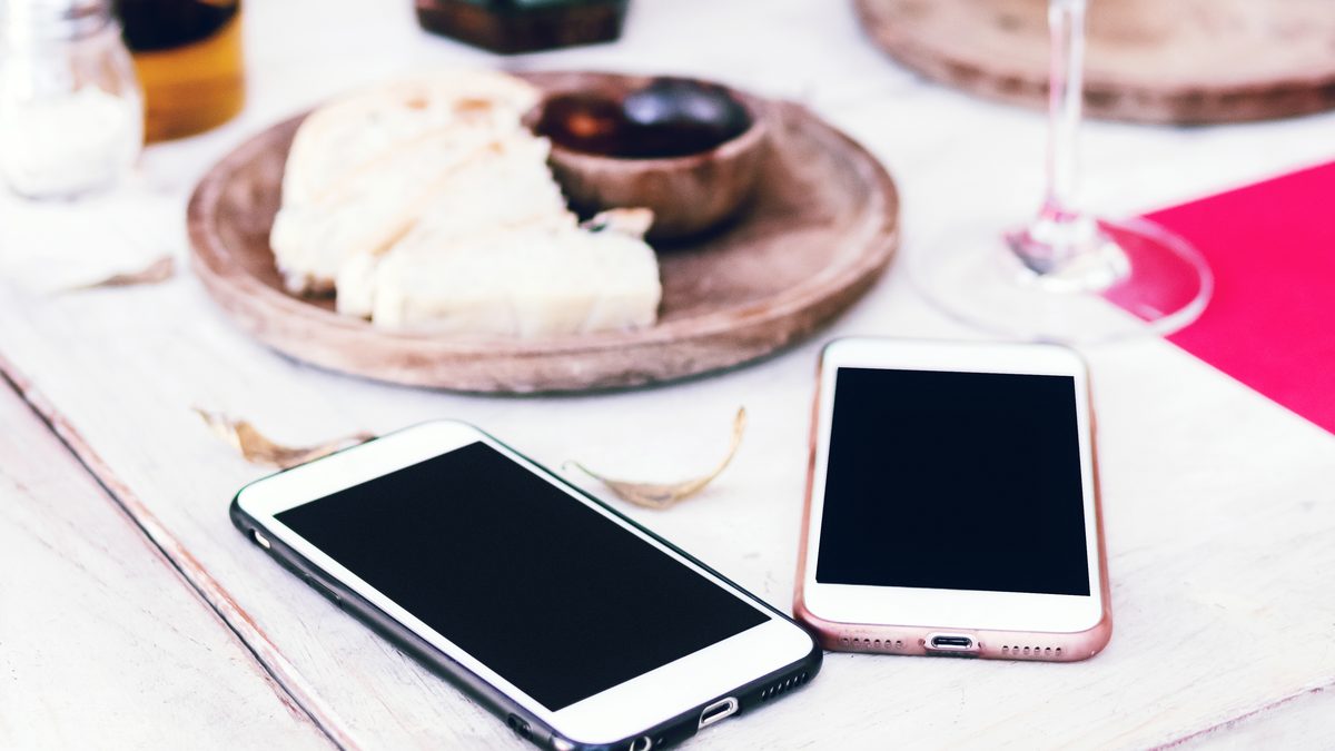 Dois celulares de tela apagada lado a lado sobre uma mesa de madeira clara, um de capa preta e outro rosé