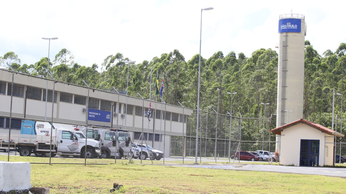 Parlatório vazio de penitenciária, com cadeiras pretas diante de guichês com vidro e interfone