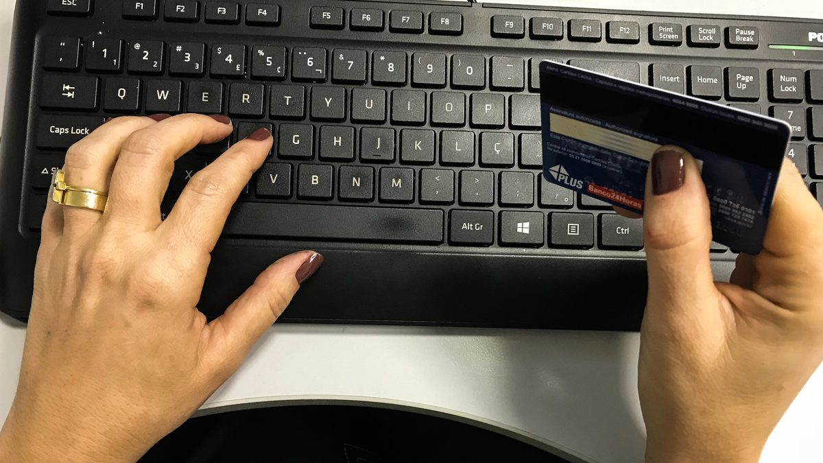 Mãos de uma mulher segurando um cartão de crédito ao lado do teclado de um computador, durante uma compra on-line