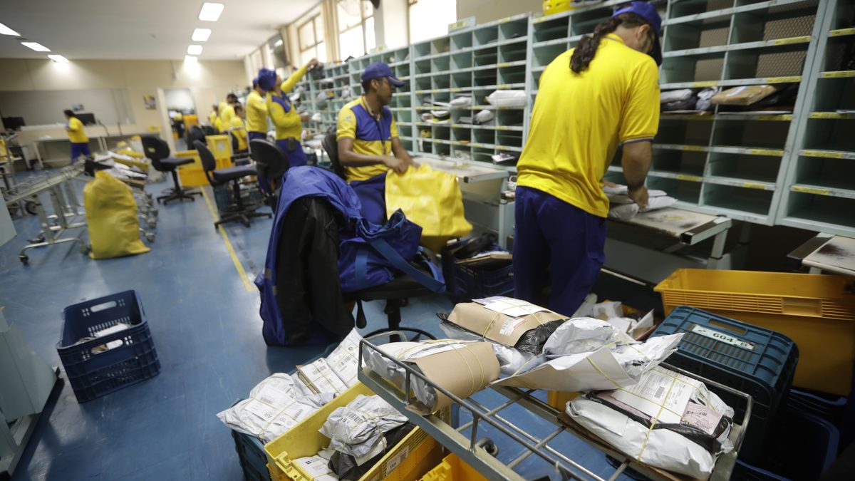 Funcionários separam encomendas em central de distribuição dos Correios em Brasília, com pacotes num carrinho em primeiro plano