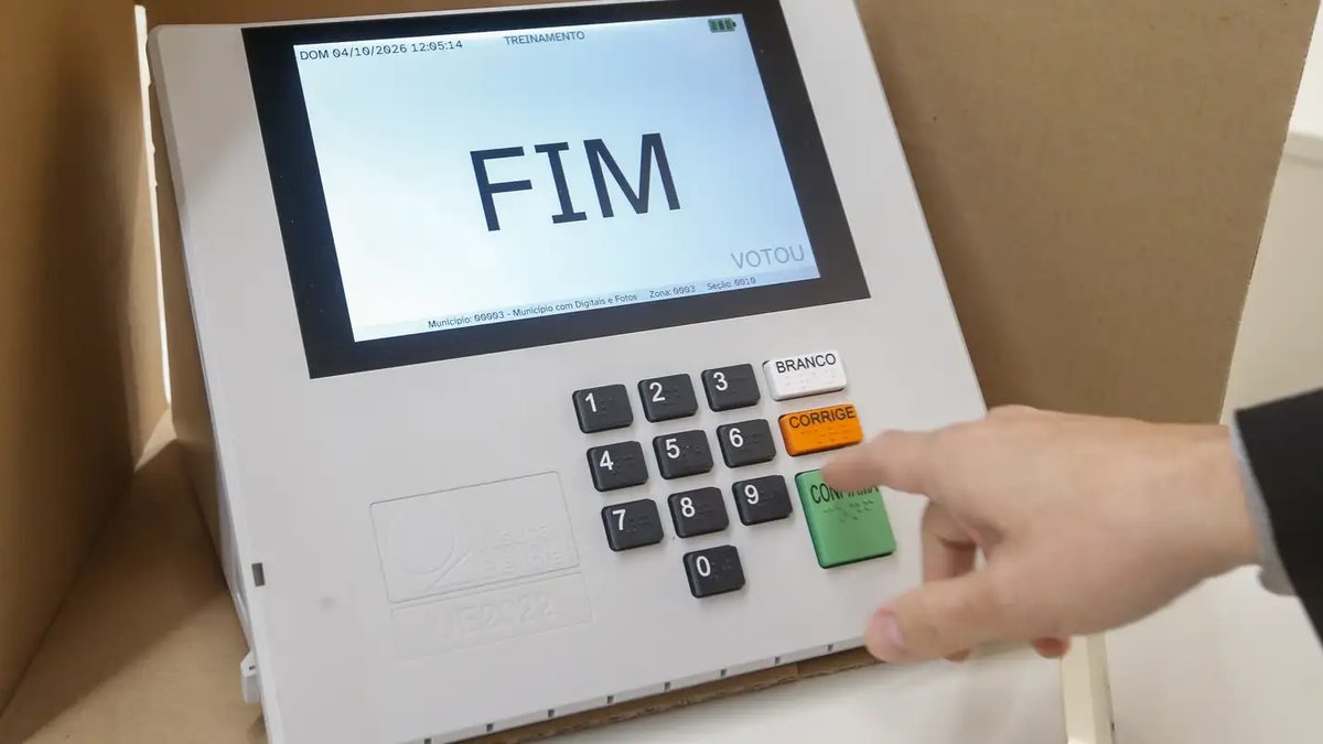 Mão aponta para o teclado de uma urna eletrônica em modo de treinamento, com a tela mostrando a palavra FIM, em demonstração do TRE-SP