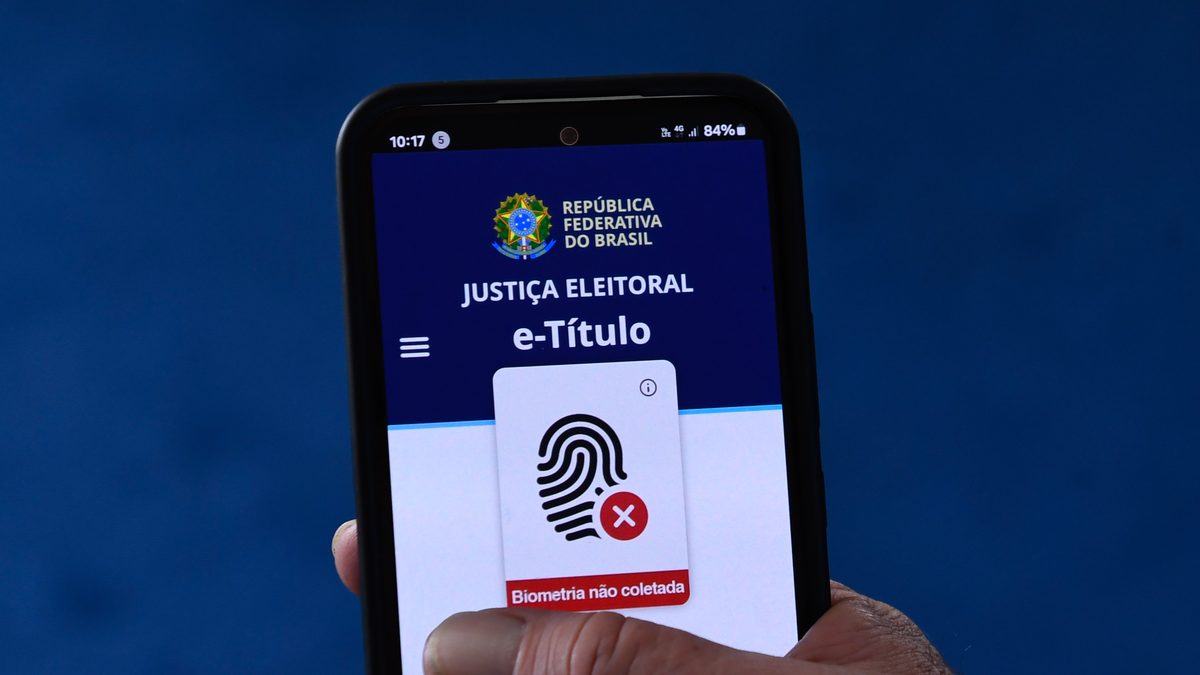 Mão de um eleitor segura um celular com a tela inicial do aplicativo e-Título, da Justiça Eleitoral, aberta, com a mensagem de biometria não coletada, sobre fundo azul