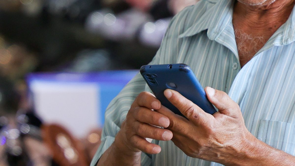 Homem idoso usando o celular, de camisa listrada, na Rodoviária do Plano Piloto de Brasília