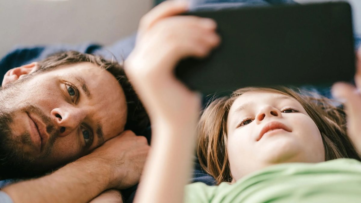 Pai deitado ao lado do filho, que usa o celular