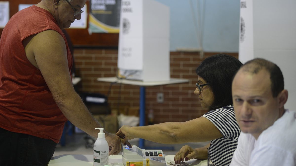 Eleitor encosta o dedo no leitor biométrico de uma seção do Colégio Itapuca, em Niterói (RJ), com cabines de votação ao fundo, no 1º turno de 2016