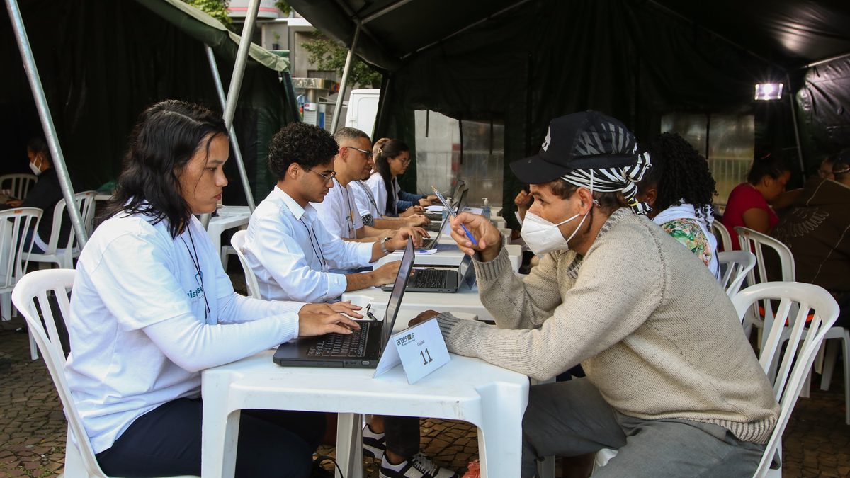 Atendentes com notebooks atendem o público em mesas sob uma tenda, no guichê 11 de um mutirão de registro civil em São Paulo