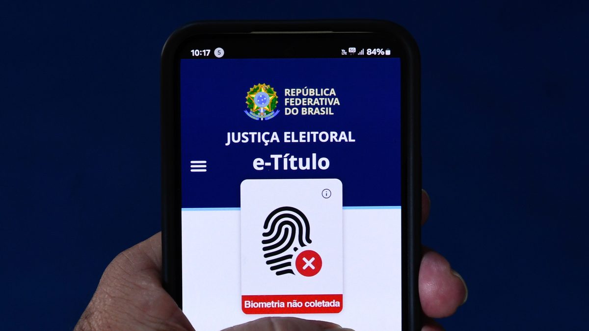 Mão segura um celular com a tela inicial do aplicativo e-Título, da Justiça Eleitoral, aberta, com o ícone de digital e o aviso de biometria não coletada, sobre fundo azul