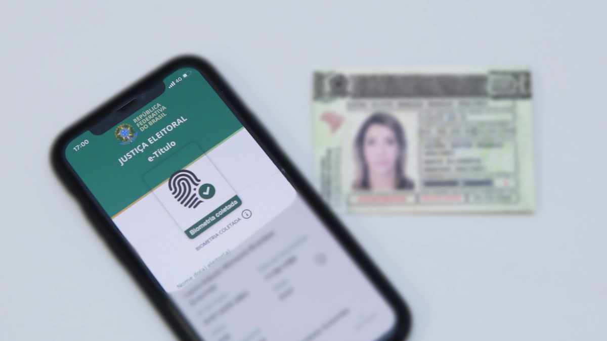 Celular com a tela do e-Título da Justiça Eleitoral mostrando o selo Biometria coletada, ao lado de um documento de identidade com foto, desfocado ao fundo