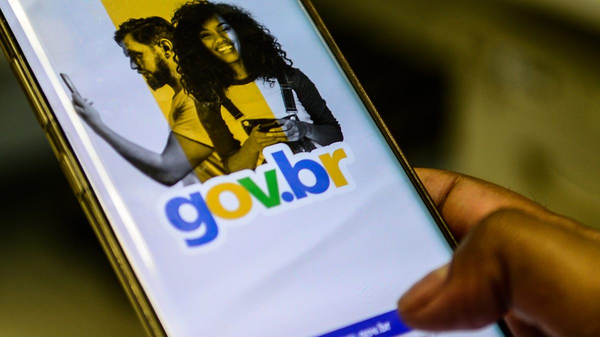 Mão segurando um celular com a tela de abertura do gov.br e o botão Entrar com gov.br