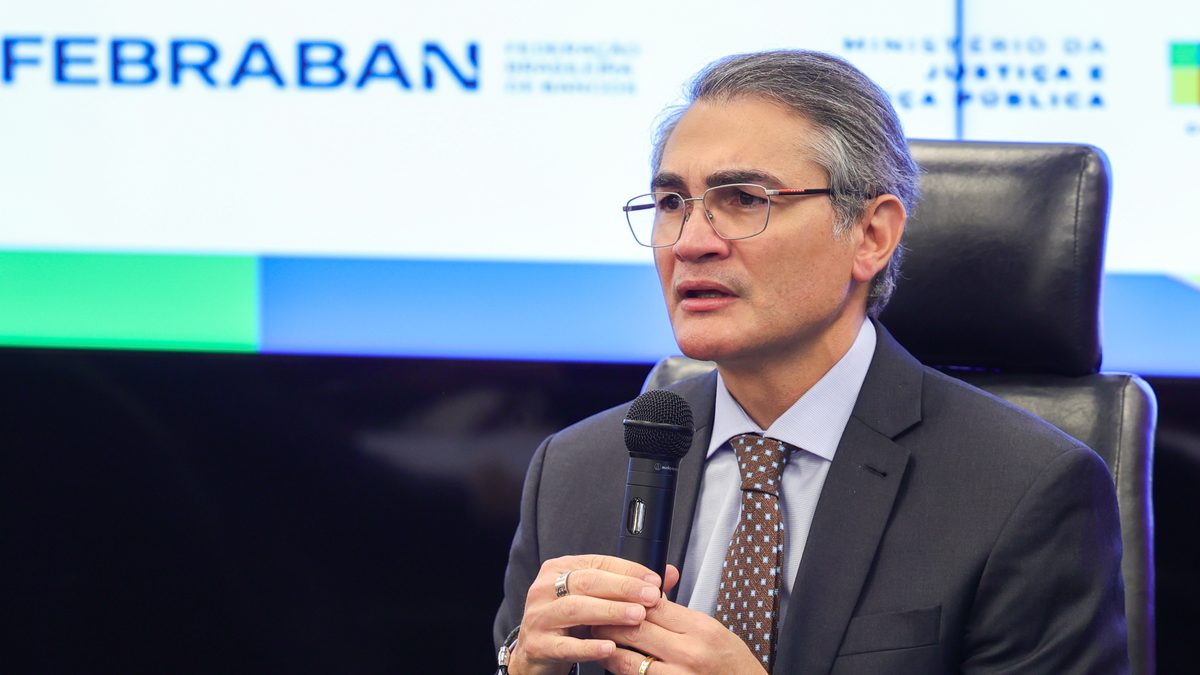 Isaac Sidney, então presidente da Febraban, fala ao microfone no lançamento do plano contra fraudes bancárias digitais, em Brasília, com a marca da Febraban no painel ao fundo