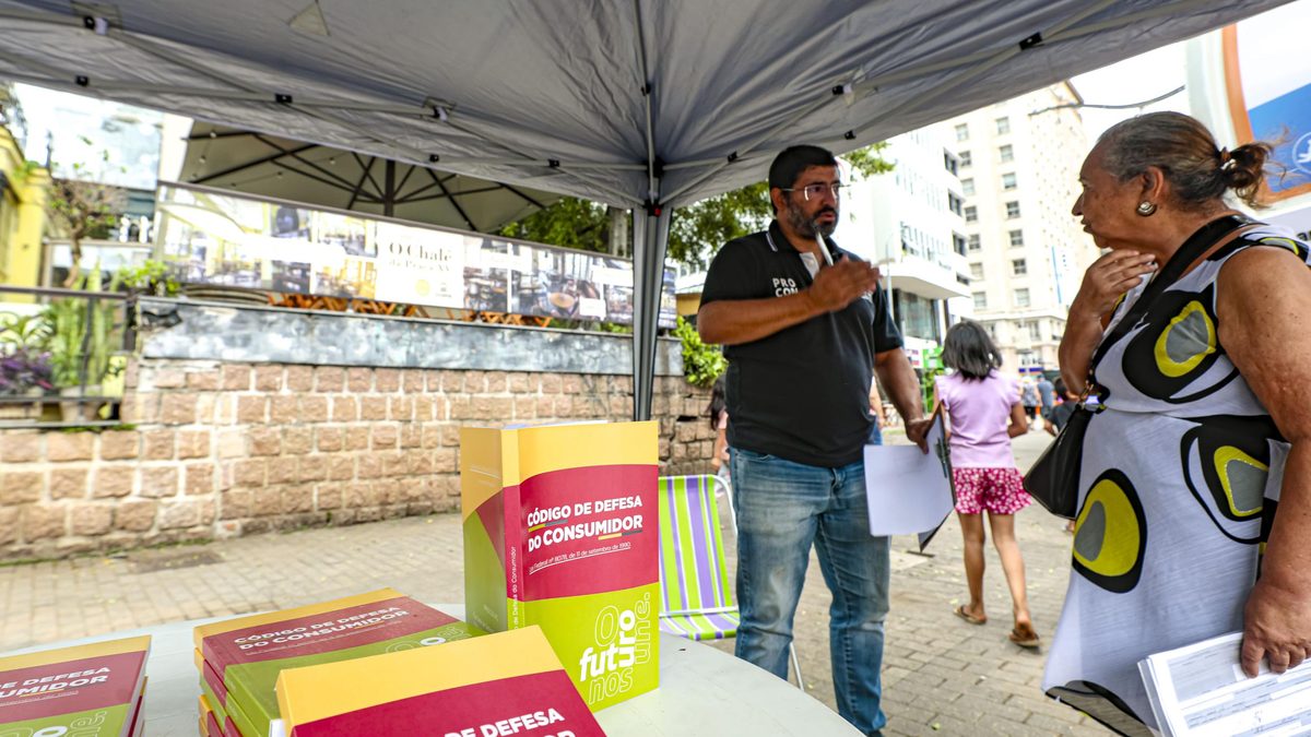 Atendente do Procon Porto Alegre conversa com uma consumidora numa tenda no Largo Glênio Peres, com exemplares do Código de Defesa do Consumidor sobre a mesa, em março de 2025