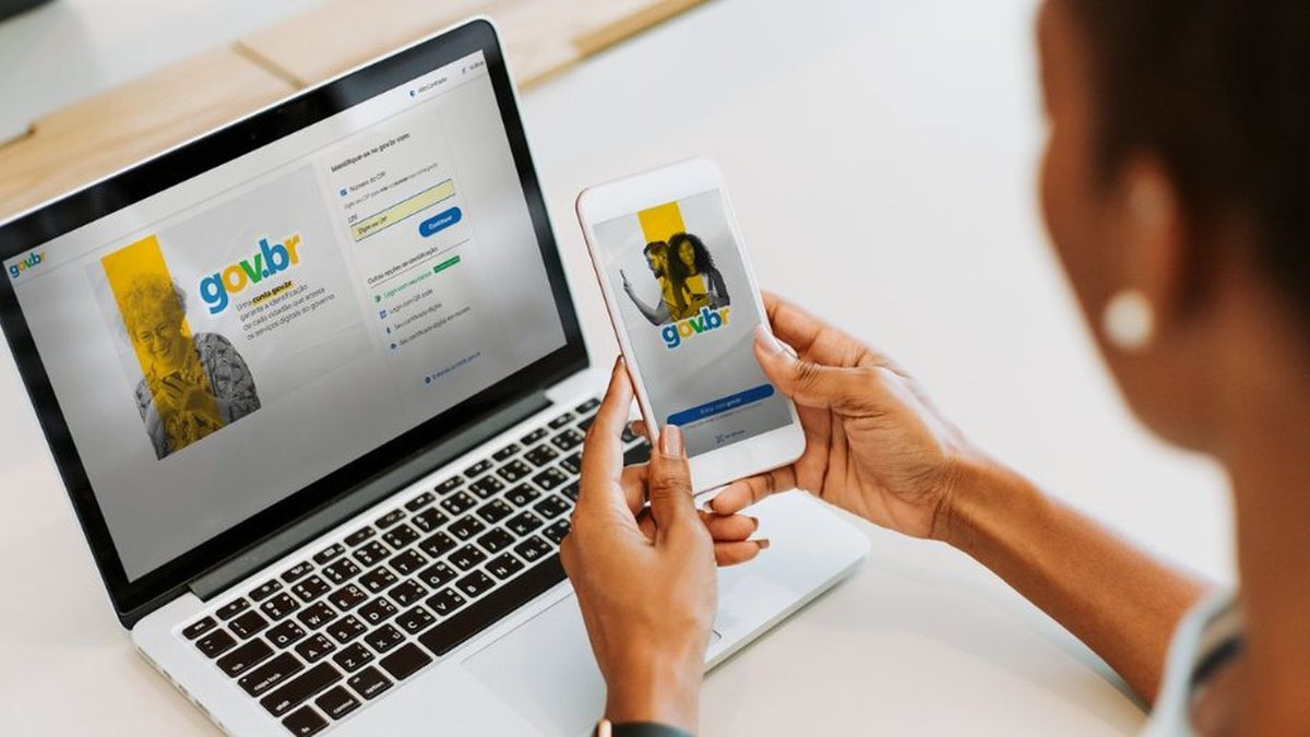 Mulher segura um celular com a tela de entrada do gov.br, diante de um notebook com a página de login do mesmo serviço, em foto de arquivo da Agência Brasil