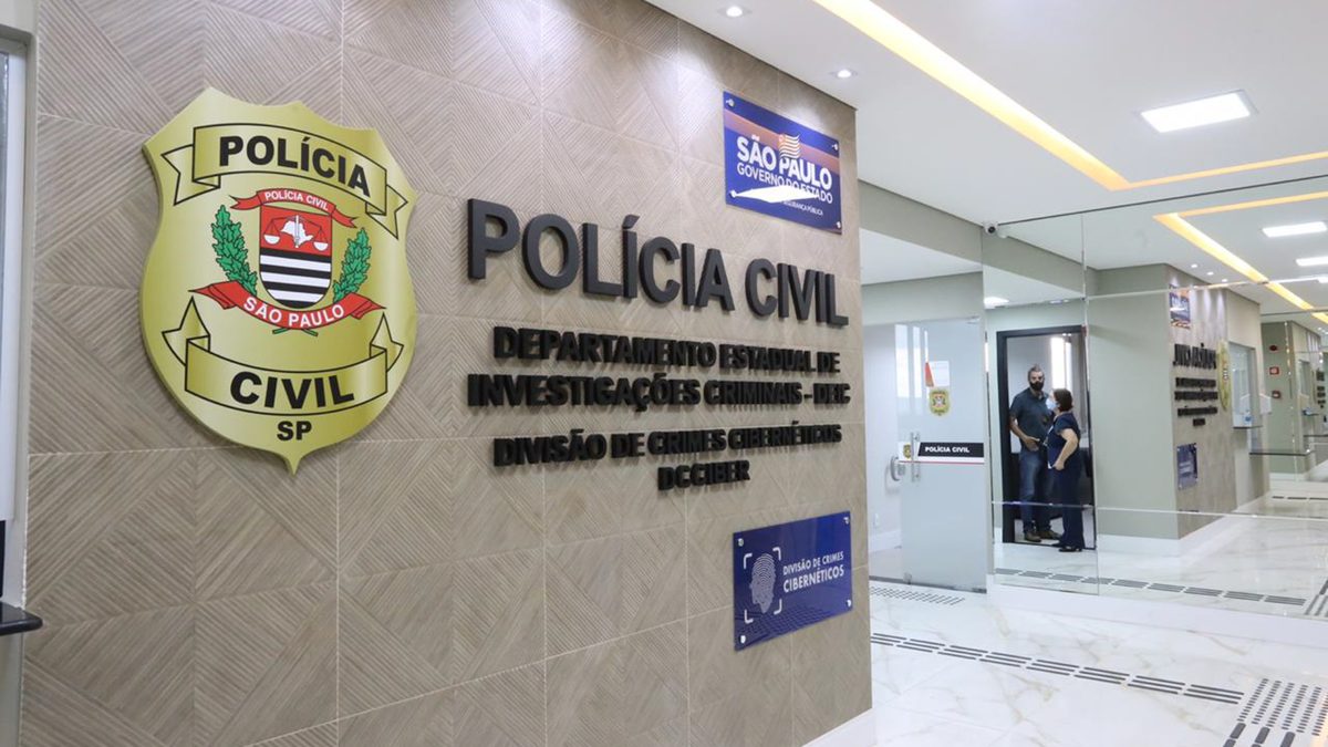 Brasão e placa da Polícia Civil de São Paulo na entrada da Divisão de Crimes Cibernéticos do Deic, com um policial à porta de uma sala ao fundo