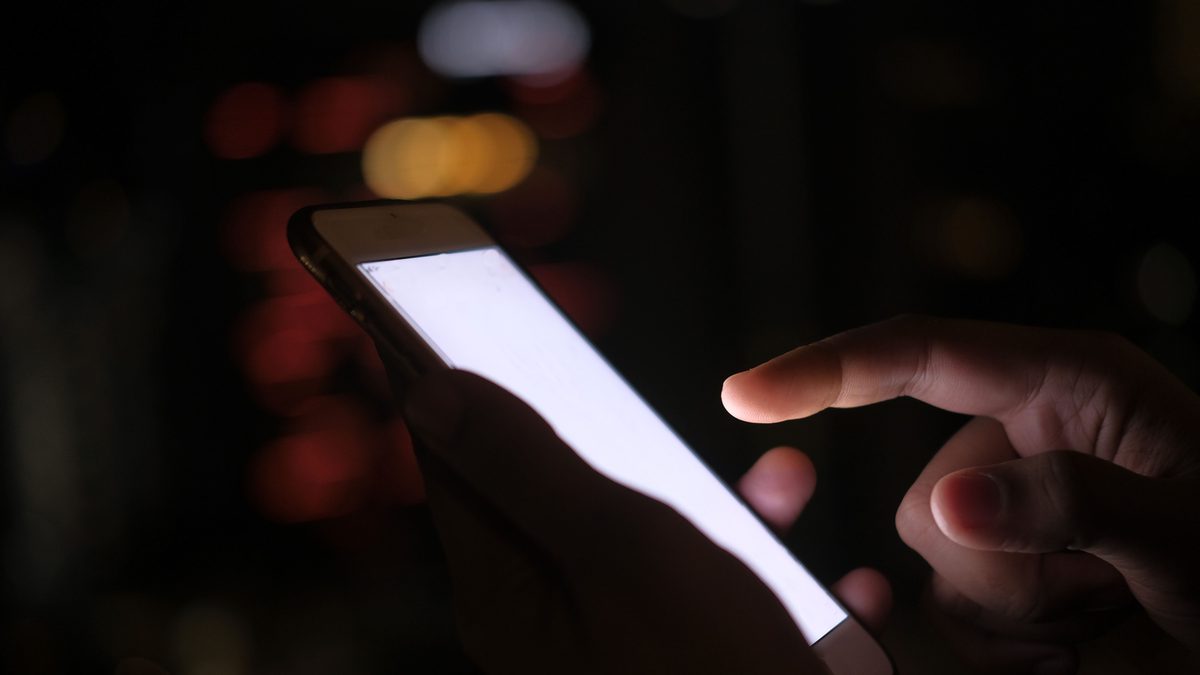 Pessoa toca com o dedo a tela luminosa de um celular no escuro
