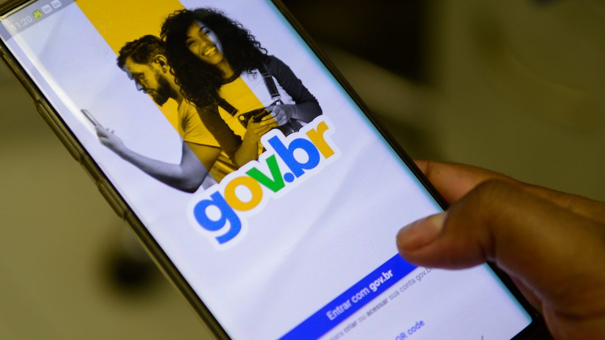 Mão segura celular com a tela de entrada do aplicativo gov.br e o botão Entrar com gov.br