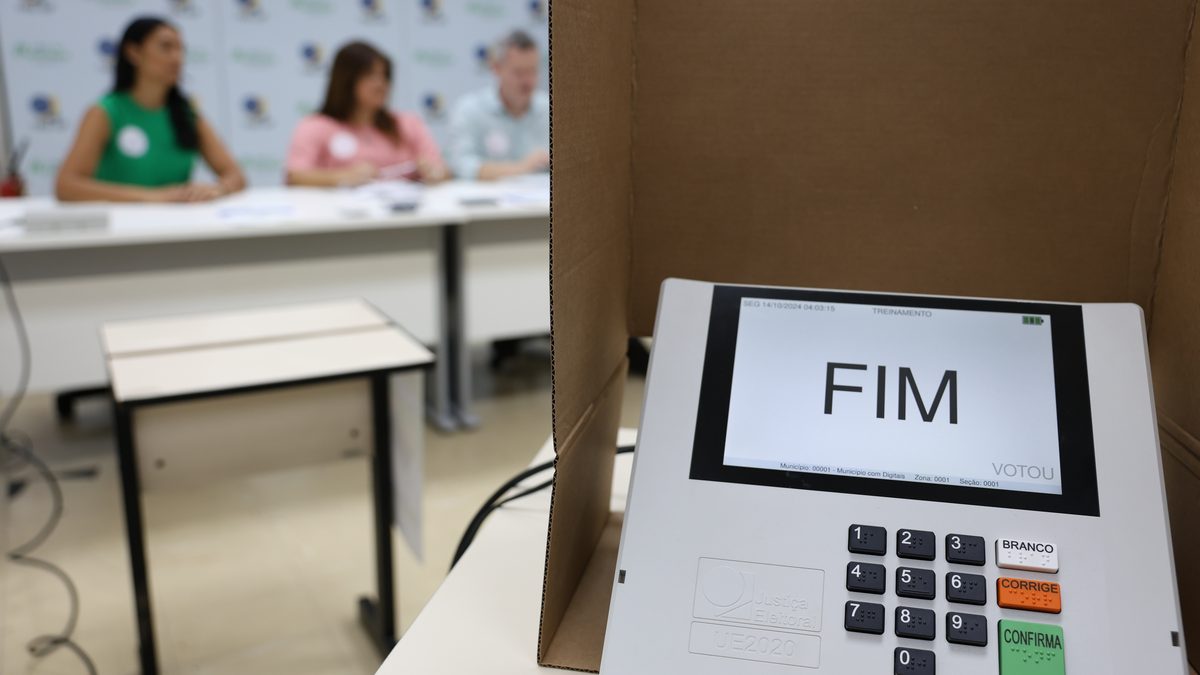 Urna eletrônica de treinamento com a tela FIM, dentro da cabine, e três mesários sentados à mesa ao fundo, em seção cenográfica do TRE-SP