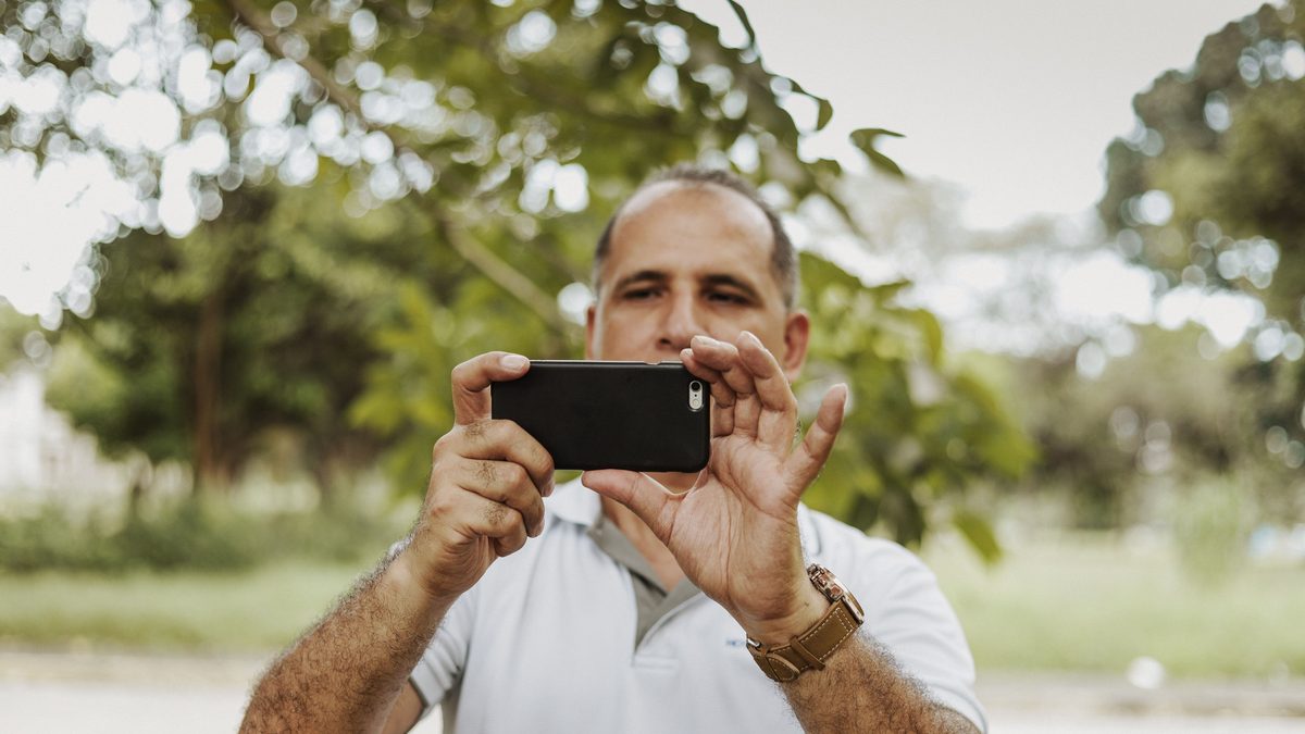 Homem de camisa polo branca fotografa com um celular na horizontal em um parque