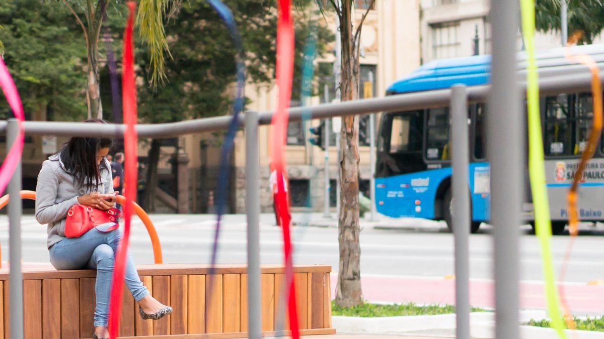 Mulher sentada num banco de madeira mexe no celular no espaço aberto da prefeitura no Largo de São Francisco, em São Paulo, com um ônibus municipal passando ao fundo