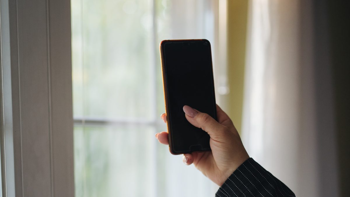Mão segurando um celular com a tela apagada diante de uma janela