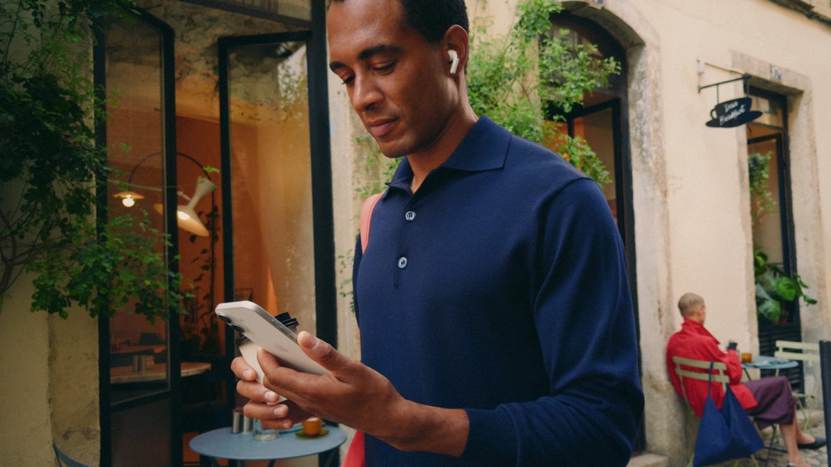 Homem de camisa azul olha para um iPhone numa rua com casas antigas e plantas, com um fone de ouvido sem fio no ouvido