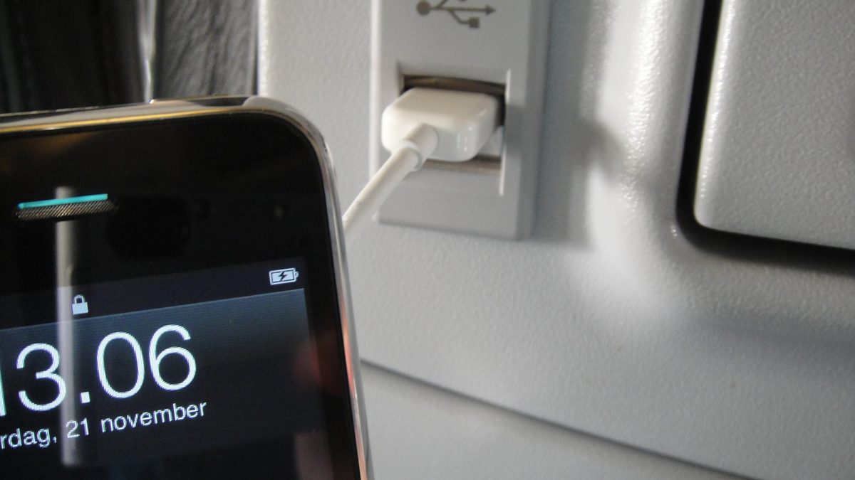 iPhone com a tela de bloqueio mostrando o ícone de carregamento, ligado por cabo a uma porta USB na poltrona de um avião