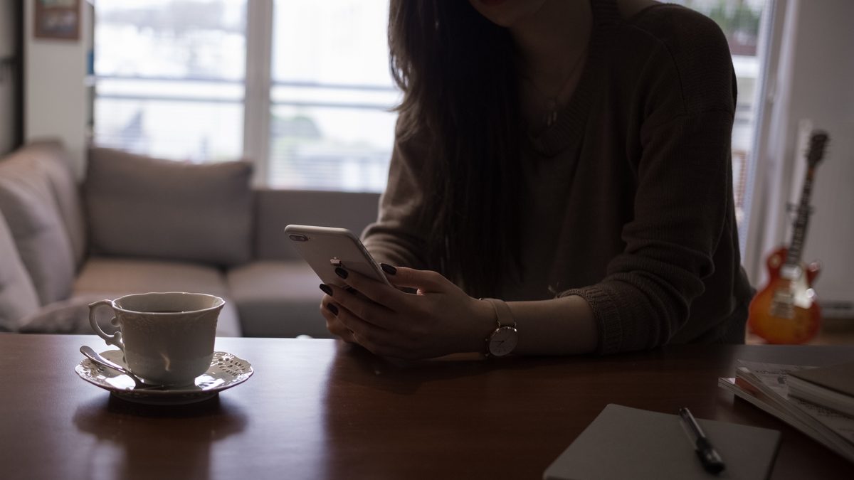 Mulher sentada à mesa segurando um iPhone com as duas mãos, ao lado de uma xícara de café