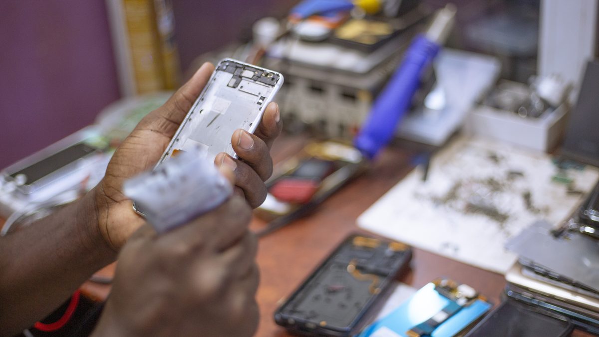 Técnico segura a carcaça aberta de um celular numa bancada de conserto com telas e aparelhos desmontados