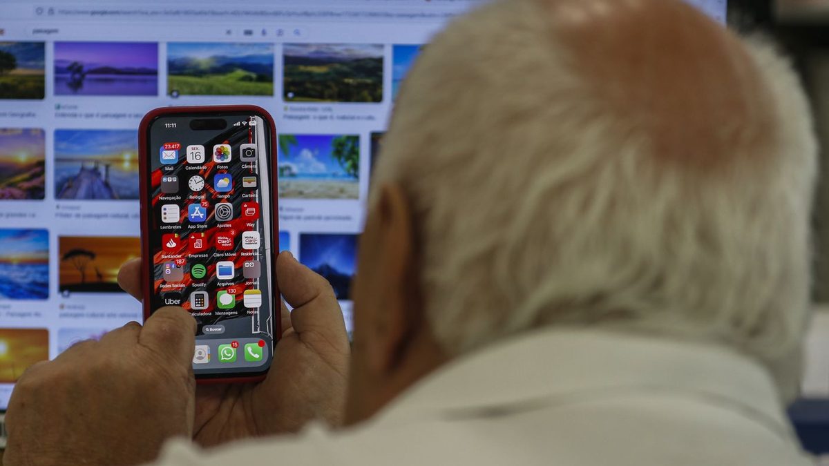 Homem idoso, visto de costas, segura um iPhone com a tela inicial aberta diante de um monitor de computador, em foto de arquivo da Agência Brasil de agosto de 2024
