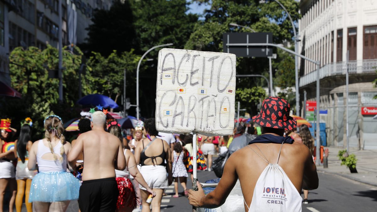 Vendedor ambulante com placa escrita Aceito Cartão e isopor de gelo caminha entre foliões em rua do centro do Rio de Janeiro, em foto de arquivo de fevereiro de 2024