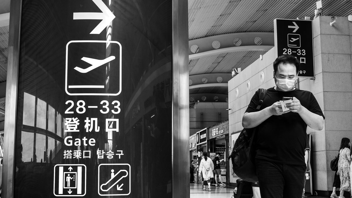 Passageiro de máscara caminha olhando o celular no saguão de embarque do antigo aeroporto internacional de Qingdao, na China, ao lado de placa com o desenho de um avião