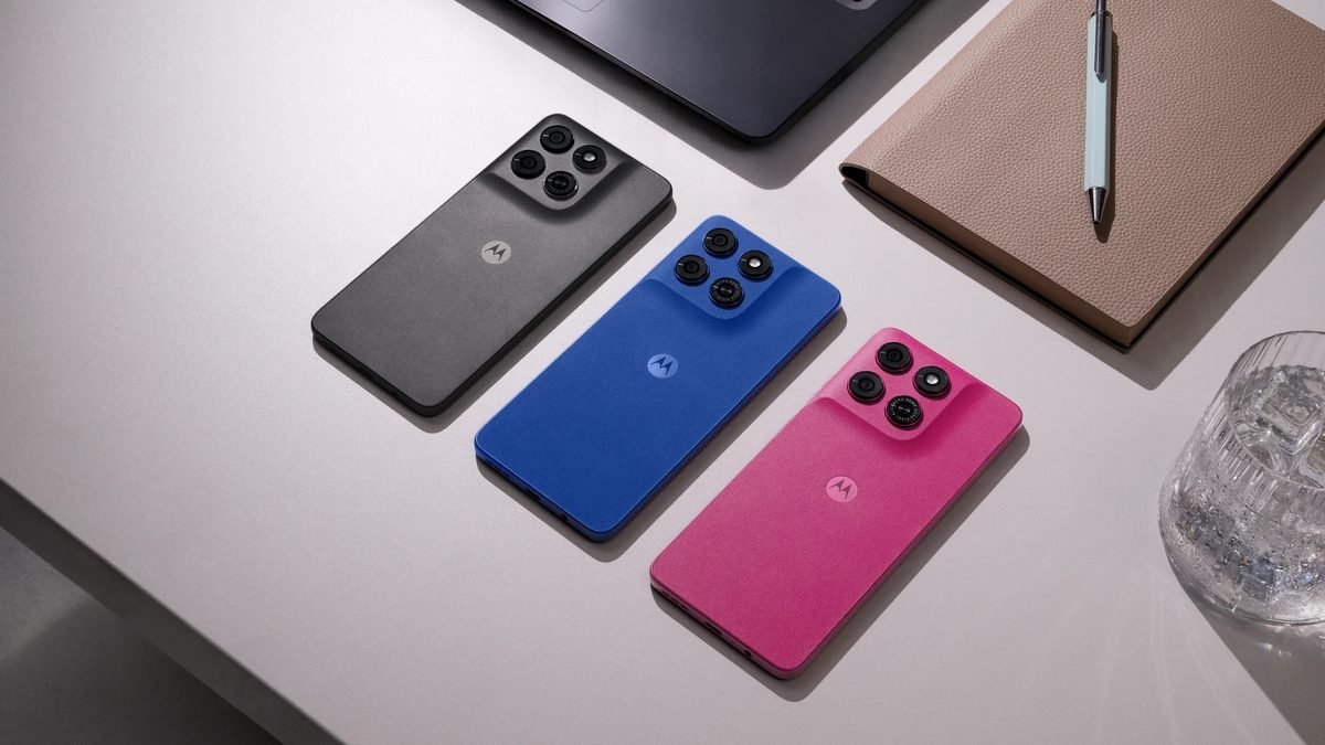 Três Moto G37 de costas sobre uma mesa clara, nas cores grafite, azul e rosa, ao lado de um notebook e de uma agenda