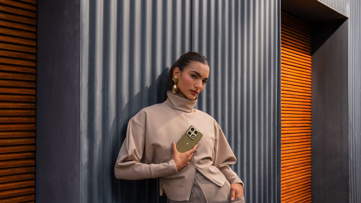 Mulher segura o Motorola Signature verde oliva junto ao peito, diante de uma parede metálica
