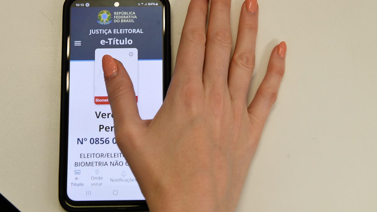 Celular com a tela inicial do aplicativo e-Título, da Justiça Eleitoral, aberta sobre uma mesa, com a mão de uma pessoa ao lado do aparelho