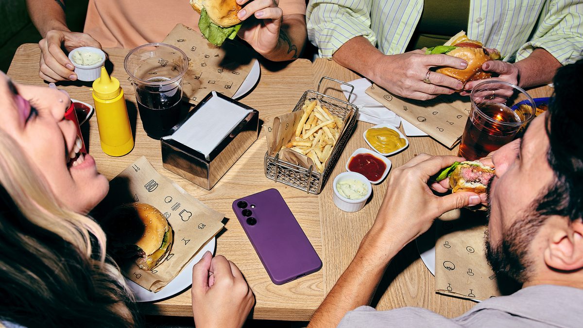 Amigos comem hambúrguer em volta de uma mesa de lanchonete, com um celular de capinha roxa ao centro, em imagem de divulgação do NuCel