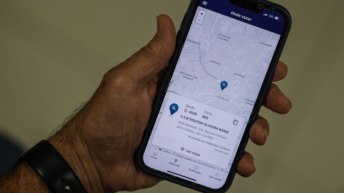 Mão segura um celular com a tela Onde votar do aplicativo e-Título, que mostra seção, zona e endereço do local de votação
