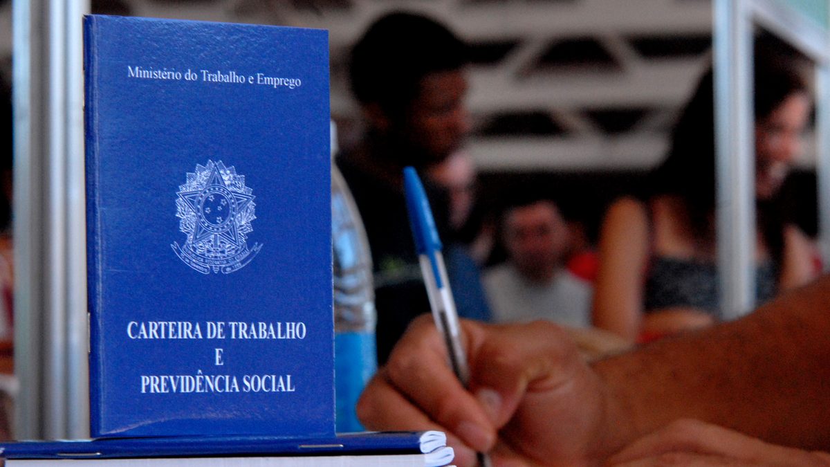 Carteiras de trabalho azuis empilhadas numa mesa da Agência do Trabalhador, em Brasília, em 2007, ao lado de uma mão que preenche um documento