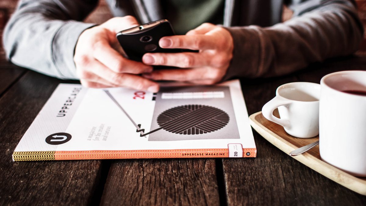 Mãos segurando um celular sobre uma mesa de madeira, ao lado de xícaras de café e de uma revista