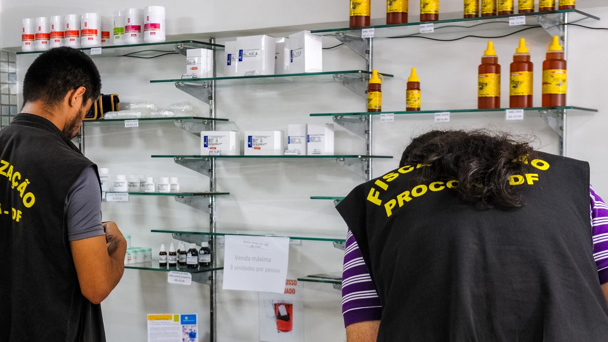 Dois agentes de fiscalização do Procon-DF, de costas, conferem prateleiras de vidro com produtos e etiquetas de preço em Brasília, em março de 2020