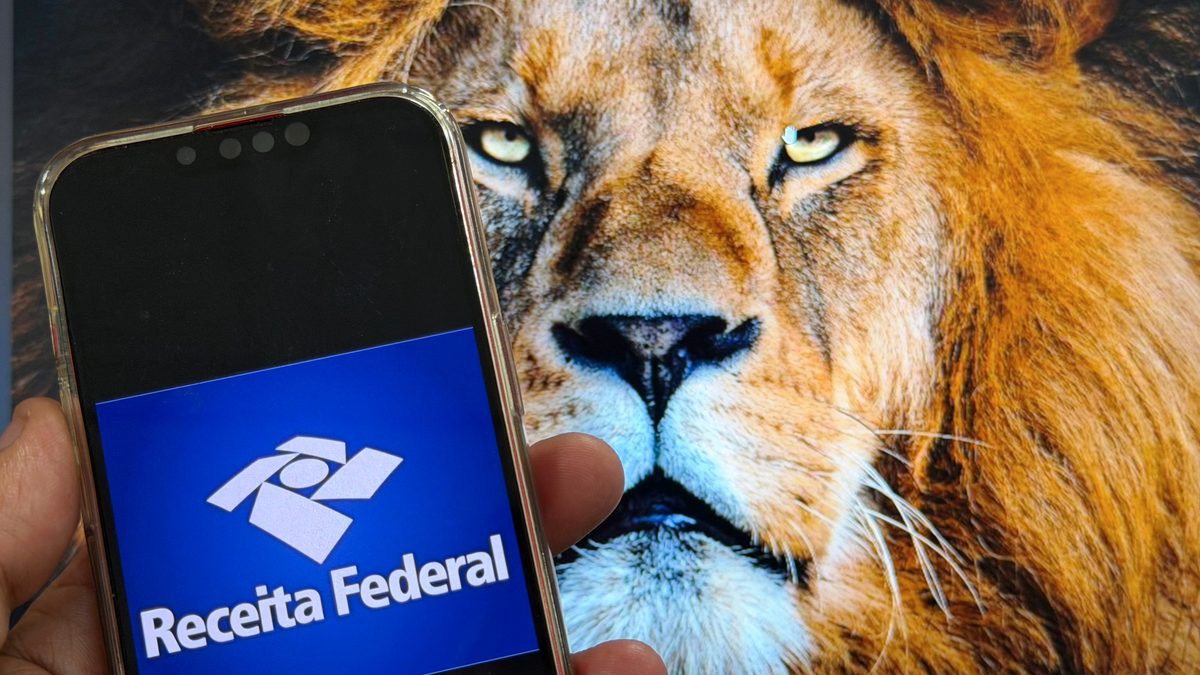 Mão segura um celular com a tela de abertura do aplicativo da Receita Federal, em frente à imagem de um leão