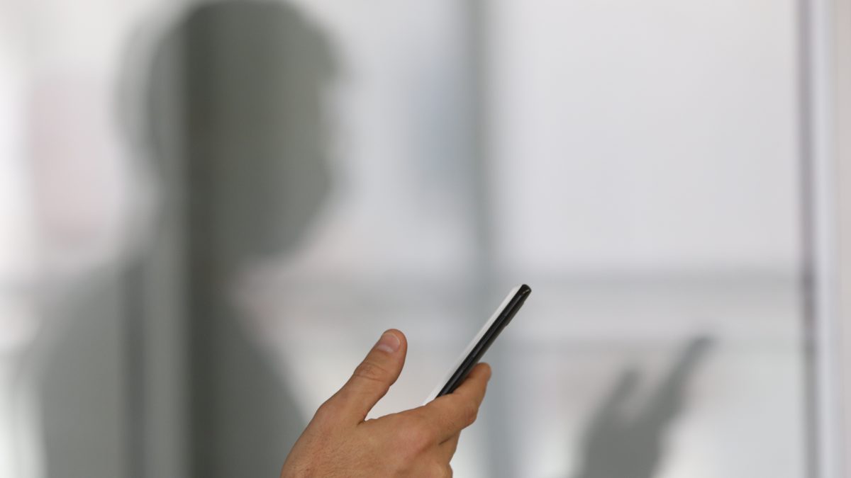 Mão segura um celular diante de uma parede clara, com a sombra de quem usa o aparelho projetada ao fundo
