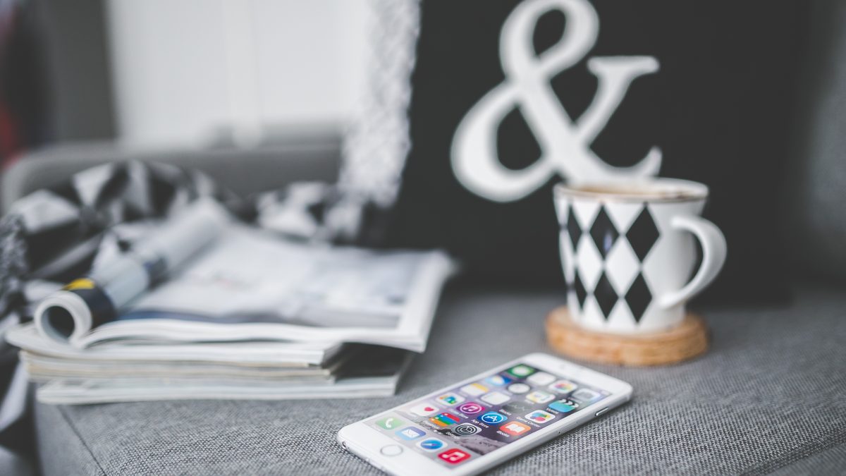 iPhone branco com a tela de apps aberta sobre um sofá cinza, ao lado de revistas e de uma xícara
