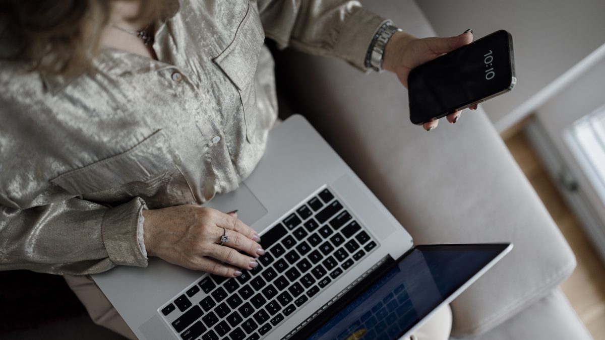 Pessoa sentada no sofá digita no notebook enquanto segura um celular na outra mão, sem o rosto aparecer
